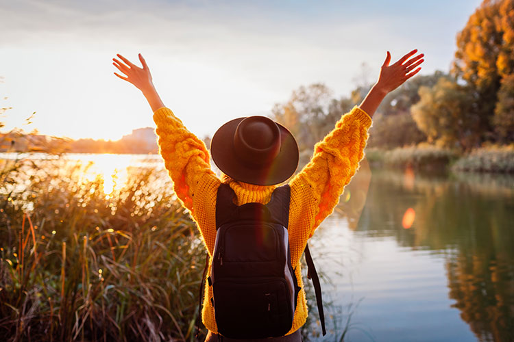 person throwing both hands up to enjoy nature while facing a pond and sunset