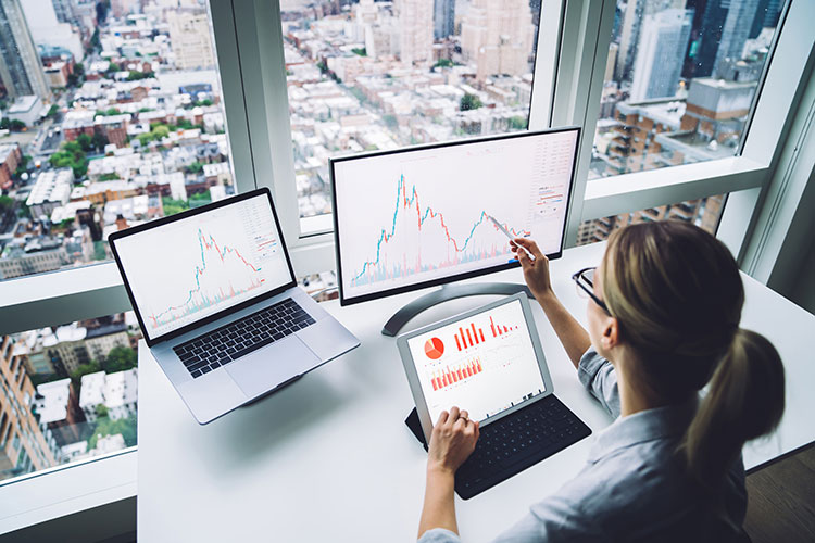 woman looking at 3 monitors with digital graphs in office overlooking the city